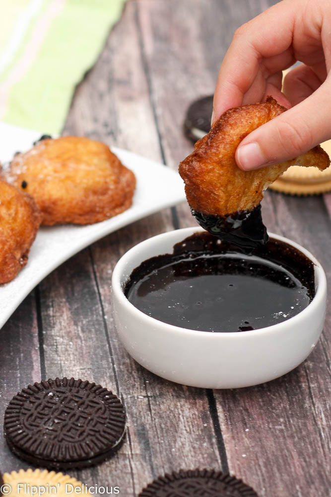 gluten free deep fried chocolate sandwich cookie being dipped in chocolate syrup with more gluten free fried oreos in the background on a white plate.