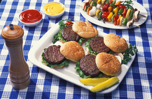 A plate of hamburgers beside vegetables on skewers, ketchup, mustard and a pepper shaker