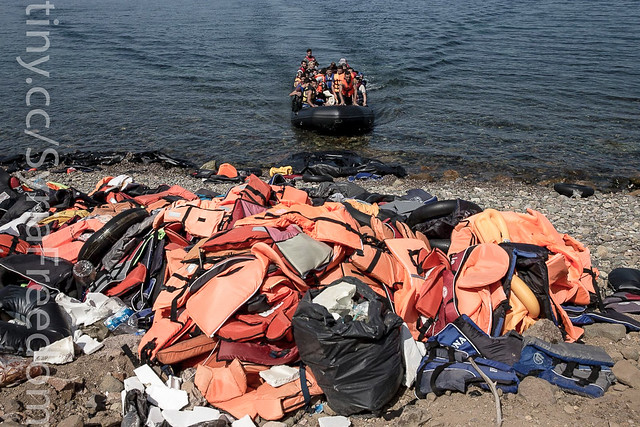 Migrants and refugees arrive by dinghy behind a huge pile of life vests after crossing from Turkey