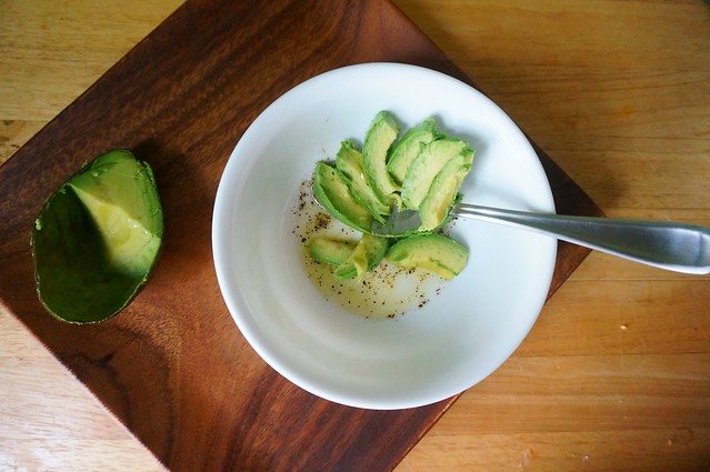 Avocado slivers have made a delicate fan on the spoon as they're scooped successively; the spoon with its avocado fan rests in a white bowl that already contains a pale yellow liquid: olive oil, lime juice, salt and pepper