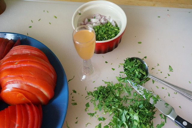 An old-fashioned cordial glass filled with amber liquid -- the vinegar dressing -- sits on a cutting board, surrounded by minced parsley. The edge of the plate of tomatoes comes in from the left, and a ramekin of minced shallot and chives sits behind.