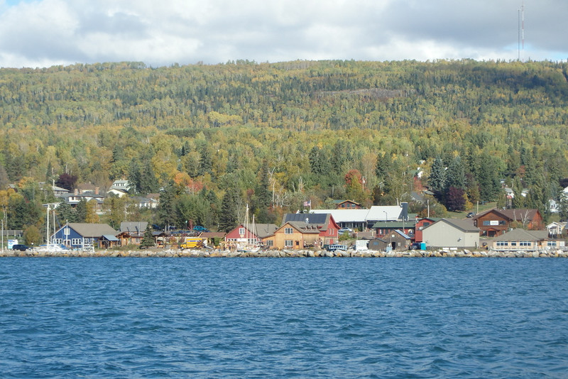 buildings next to big rocks along the shore, lake in the foreground, mountain with fall trees in the background