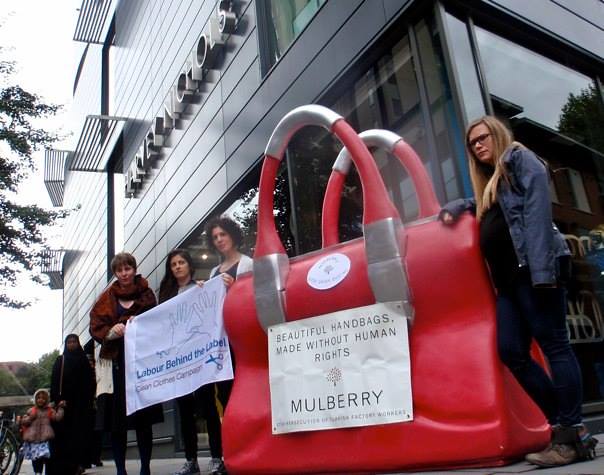 Protesters stand next to a huge purse labeled "Mulberry" and give flyers to passersby