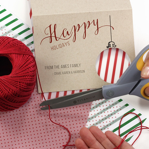 scissors cutting red twine placed next to printed holiday card and decorative papers