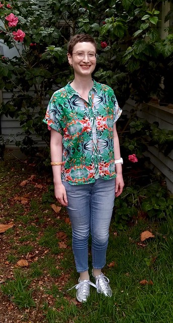 A woman stands in front of hibiscus plant. She wears a green, tropical print shirt, skinny jeans and silver runners.