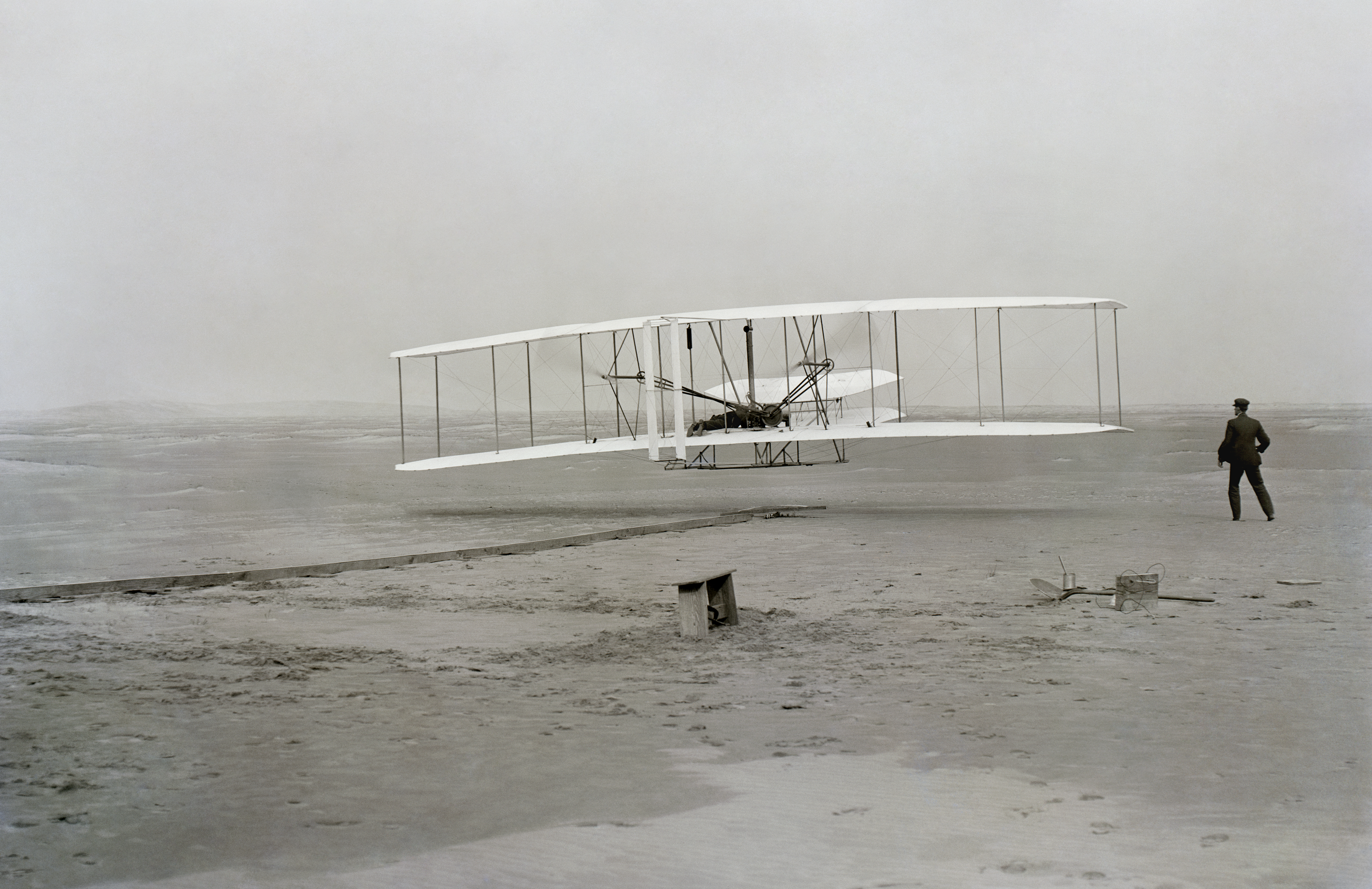 First successful flight of the Wright Flyer, by the Wright brothers. The machine traveled 120 ft (36.6 m) in 12 seconds at 10:35 a.m. at Kill Devil Hills, North Carolina. Orville Wright was at the controls of the machine, lying prone on the lower wing with his hips in the cradle which operated the wing-warping mechanism. Wilbur Wright ran alongside to balance the machine, and just released his hold on the forward upright of the right wing in the photo. The starting rail, the wing-rest, a coil box, and other items needed for flight preparation are visible behind the machine. This is described as 