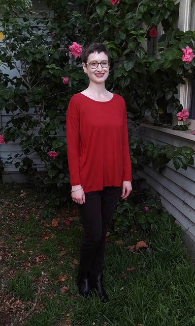 A woman stands in front of a hibiscus plant. She wears a drop shoulder, long sleeve red tee and black skinny pants.