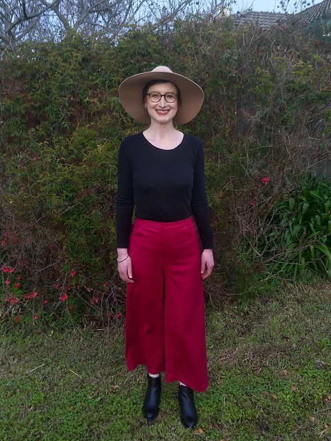 A woman stands against a garden fence. She wears magenta wide leg crop pants, a black skivvy, black ankle boots, and a tan felt hat.