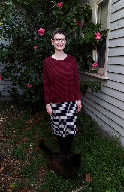 A woman stands in front of a hibiscus plant. She wears a burgundy drop shoulder, long sleeve knit tee with blue white stripe skirt. A cat walks around her.