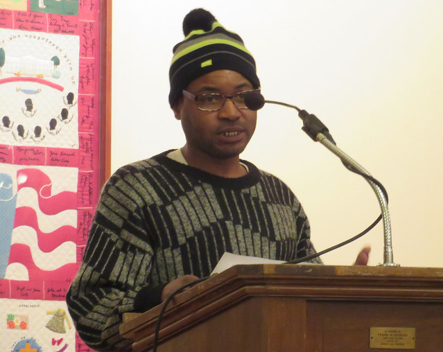 A man in a sweater and winter hat speaks at a podium.