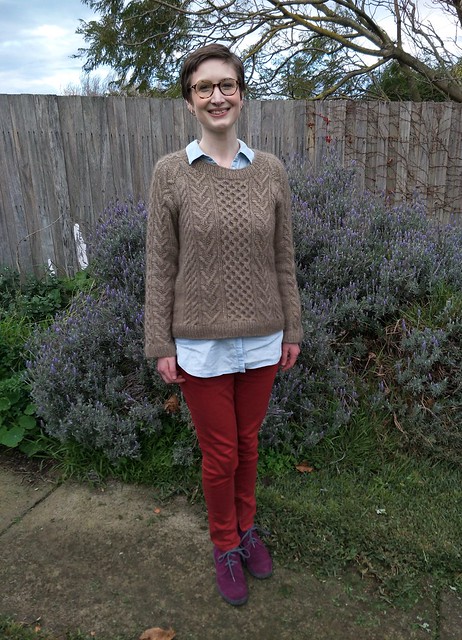A woman stands in front of a garden fence. She wears a handknit cabled jumper, denim shirt, red jeans and purple suede boots. She is smiling.