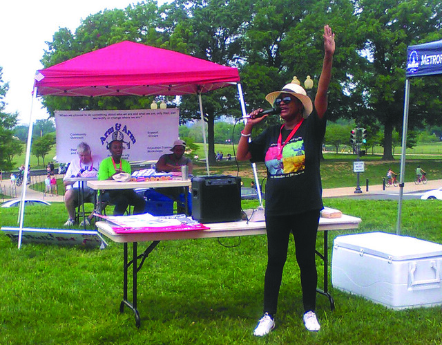 Photo of a woman holding her hand in the air and speaking into a microphone. She is wearing a wide-brimmed hat and sunglasses, standing next to a table and a cooler. Several other people sit at a table behind her, under a canopy.